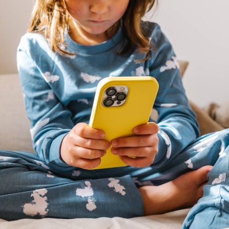 Ein Kleinkind im Schlafanzug sitzt auf einem Bett und klickt auf einem Smartphone in gelber Hülle.
