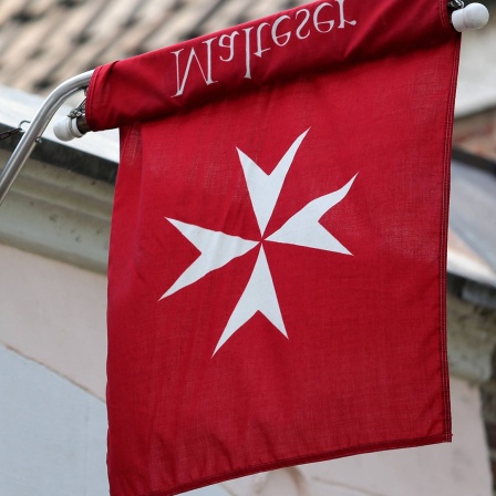 Eine Flagge mit dem rot-weißem Logo der Malteser hängt seitlich an einer Außenwand.