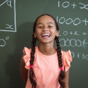 Porträt eines lachenden Schulmädchens. Es steht vor einer Kreidetafel mit Matheaufgaben. 