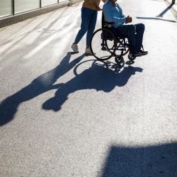 Eine Person schiebt eine andere Person in einem Rollstuhl. Ihr Schatten ist auf der Strasse sichtbar.