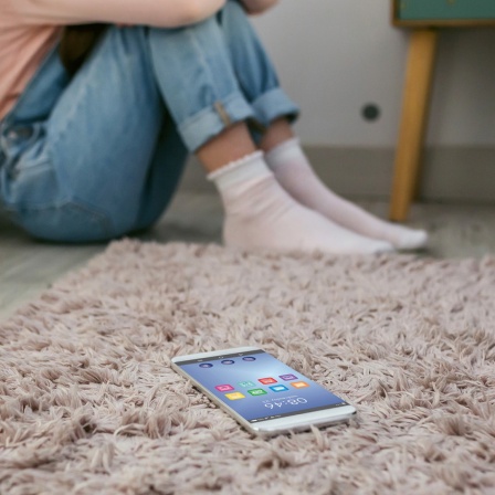 Symbolfoto: Eine junge Frau hockt in einer Zimmerecke, auf dem Boden liegt ein Smartphone.