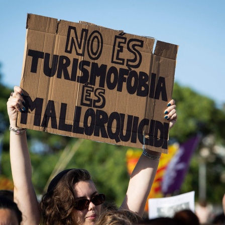 Junge Frau auf einer Demo gegen Massentourismus auf Mallorca hält ein Schild mit der Aufschrift ""no és turismofobia, és mallorquicidi" (Deutsch: "Es ist keine Tourismusfeindlichkeit. Es ist Mallorquinermord.") 