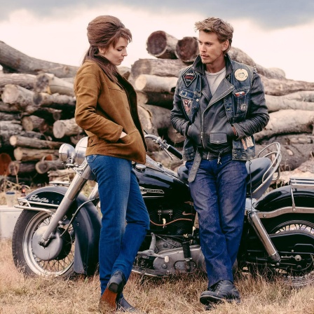 Szene aus dem Film "The Bikeriders" von Jeff Nichols, 2024. Die Schauspieler Austin Butler und Jodie Comer stehen vor einem Motorrad.