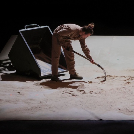 Zwei Personen arbeiten auf einem sandigen Boden mit Werkzeugen.
