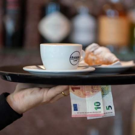 Nahaufnahme eines Arms eines Kellners oder einer Kellnerin in einem schwarzen Pullover, der ein Tablett mit einer Tasse und einem Croissant hält – außerdem befinden sich ein 5 Euro Schein und ein 10 Euro Schein in der Hand unter dem Tablett. 
