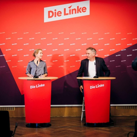 Jan van Aken und Ines Schwerdtner, Vorsitzenden der Partei Die Linke, blicken sich bei einer Pressekonferenz an. Im Hintergrund sieht man den Schriftzug "Die Linke".