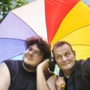 Devid Striesow und Axel Ranisch lehnen sich unter einem bunten Regenschirm aneinander und lauschen den Tönen aus einem Kopfhörer.