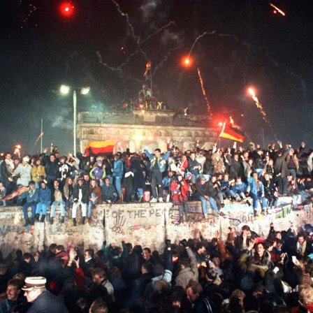 Zahlreiche Menschen sitzen auf und stehen vor der Berliner Mauer, im Hintergrund ist das Brandenburger Tor zu sehen, Pyrotechnik leuchtet in der Luft.