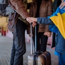 Drei Personen stehen mit Gepäck an einem Bahnhof. Auf einer Jacke ist eine ukrainische Flagge zu sehen.