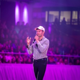 Patrick Thomalla steht in einer Arena und spricht in ein Mikrofon. Er trägt schwarze Reiterhosen und ein Basecap.