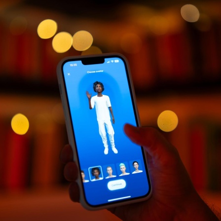 Eine Hand hält ein Smartphone auf dem eine virtuelle Person zu sehen ist, die in die Kamera blickt. 