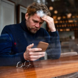 Mann mit lockigem Haare sitzt an Fenster-Tresen eines Cafés, schaut auf Smartphone und greift sich sorgenvoll am Kopf. Daneben liegt dratige Brille. Ansicht von außen durch Scheibe. Leichte unscharge Spiegelungen in Scheibe, unscharfer Hintergrund.