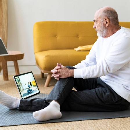 Sport vom Sofa aus: Um fit zu werden, muss man nicht in teure Studios gehen. Findet auch der ältere Mann auf dem Foto, der im bequemen Yoga-Outfit auf einen Laptop-Bildschirm schaut. 