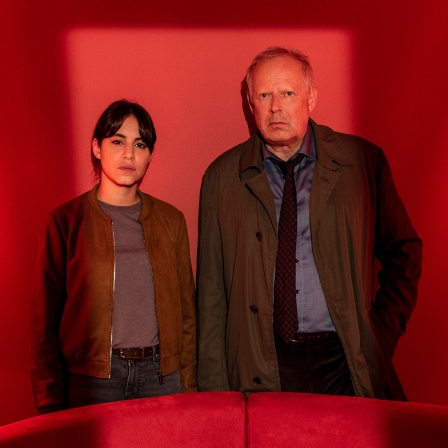 Almila Bagriacik und Axel Milberg stehen bei einer Szene im Tatort "Borowski und das hungrige Herz" hinter einem roten Sofa