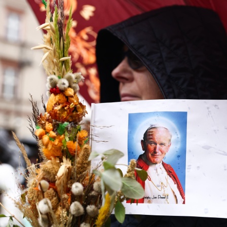 Eine Person mit Sonnenbrille und schwarzer Kapuze trägt eine Illustration von Papst Johannes Paul II. und einen Strauss mit Trockenblumen während einer Demonstration in Krakow.