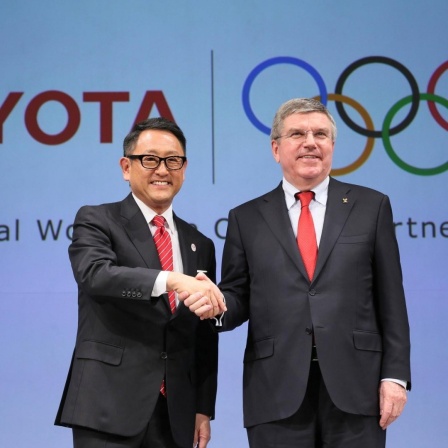 Toyota-Konzernchef Akio Toyoda und IOC-Präsident Thomas Bach schütteln sich die Hände, im Hintergrund die Logos von Toyota und den Olympischen Spielen
