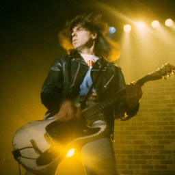 Johnny Ramone von den Ramones live beim SuperBang '93 in der Eilenriedenhalle. Hannover, 12.12.1993. Er spielt dymanisch die Gitarre, seine Haare fliegen im Wind. Die Bühen ist in gelbes Licht geflutet und leicht nebelig. 