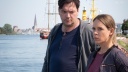 Eine Frau und ein Mann stehen an einem Ufer vor Schiffen und blicken ernst.