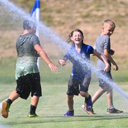 Eine Gruppe von fußballspielenden Jungen erfrischt sich an einem heißen Sommertag auf dem Spielfeld mit Wasser aus einem Rasensprenger.
