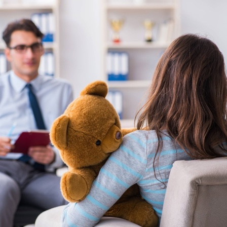 Eine Frau mit einem Teddybär im Ar sitzt auf einer Couch und spricht und ihr gegenüber sitzt ein man und macht sich Notizen.
