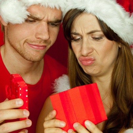Ein Mann und eine Frau mit Weihnachtsmützen blicken unzufrieden auf ein Geschenk.