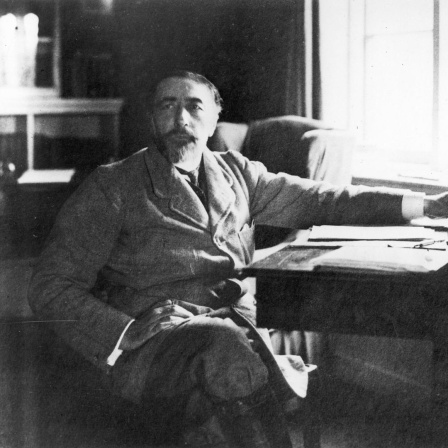 Der Schriftsteller Joseph Conrad (1857-1924) sitzt mit übergeschlagenen Beinen seitlich an einem Schreibtisch.