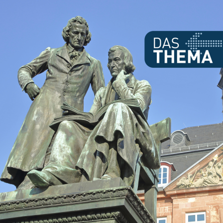  eine Statue zweier Männer, die in historische Kleidung gekleidet sind. Einer sitzt und liest in einem Buch, während der andere stehend auf das Buch schaut. Es handelt sich um das bekannte Brüder-Grimm-Denkmal in Hanau, das an die berühmten Sprachwissenschaftler und Märchensammler Jacob und Wilhelm Grimm erinnert.