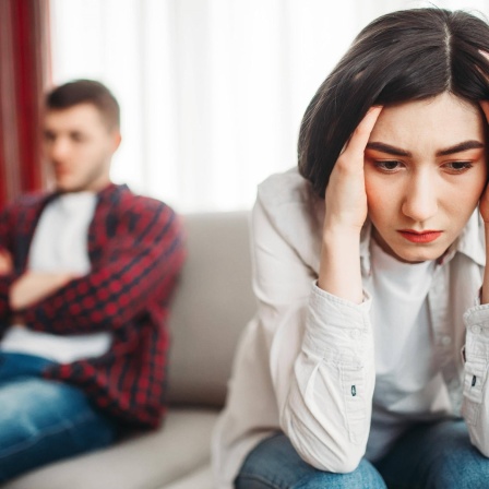 Eine Fraue rauft sich die Haare, im Hintergrund ist verschwommen ein Mann zu erkennen, der auf einer Couch sitzt. 