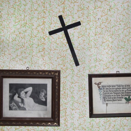 Ein schiefes Kreuz und zwei alte Bilder hängen an einer Wand.