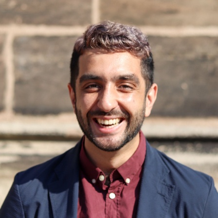 Tareq Sydiq, Politikwissenschaftler, steht vor einer Steinmauer in der Sonne und lacht.