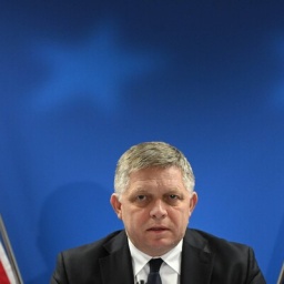 Robert Fico sitzt zwischen der slowakischen und der europäischen Flagge und schaut in die Kamera