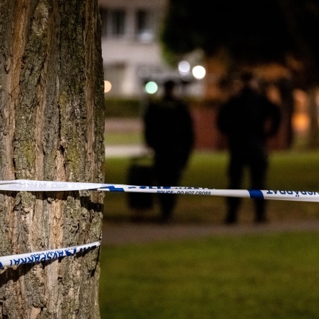 Polizeiabsperrband um einen Baum gewickelt, im Hintergrund zeichnen sich dunkel gekleidete Menschen ab.