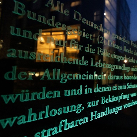 Textauszug des Grundgesetzes auf einer Glaswand, im Hintergrund der Deutsche Reichstag mit einer Deutschland-Fahne in der Dämmerung.
