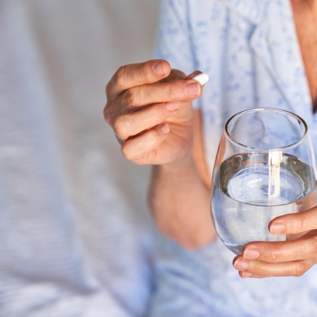 Eine ältere Frau hält eine Tablette und ein Wasserglas in der Hand, während sie im Schlafanzug auf einem Krankenhausbett sitzt.