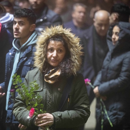 Eine Frau steht im Vordergrund mit einer Blume in der Hand, hinter ihr stehen weitere Menschen.