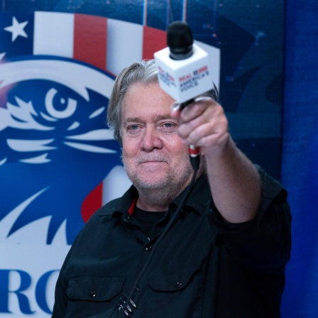 Steve Bannon hält ein Mikrofon hoch während seiner Radiosendung auf der Conservative Political Action Conference, CPAC 2023. Im Hintergrund eine Flagge der USA mit Adler.
