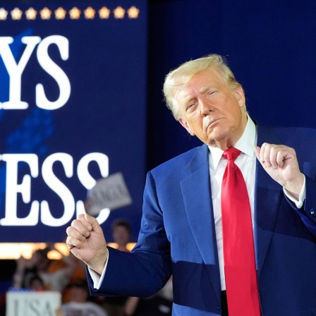 Donald Trump hat die Augen geschlossen und scheint vor einen Schild, auf dem "100 Days of Greantness" steht, zu tanzen. 