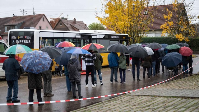 Menschen warten mit Regenschirmen in einer Schlange vor einem Impfbus.
