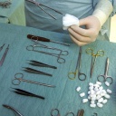 In einem OP-Saal steckt ein Arzt an einem Tisch mit Operationsbesteck mit einer Hand, die in einem sterilen Gummihandschuh steckt, einen Tupfer in eine Zange. 