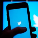 Ein Hand hält ein Smartphone auf dessen Bildschirm das Twitter-Symbol zu sehen ist, das auch im Hintergrund erscheint