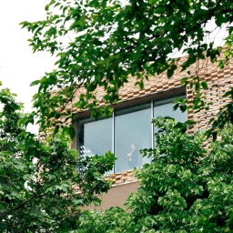 Die Ehrlichs haben sich ihr Traumhaus gebaut – es gibt Komplikationen. Zu sehen: Bäume im Vordergrund, dahinter der Teil eines modernen Hauses mit großen Fenstern. Im Fenster steht eine Frau, die nach draußen blickt.