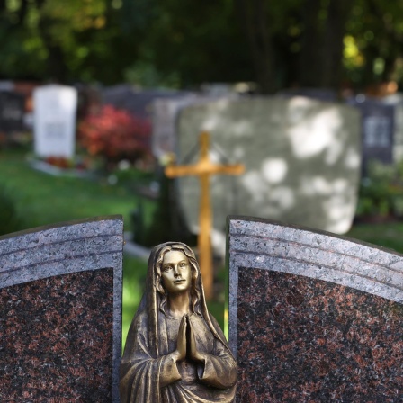 Eine Engelsfigur steht auf einem Friedhof vor Grabsteinen. 