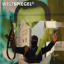 Ein vermummter Kämpfer steht mit ausgebreiteten Armen und einem Maschinengewehr in der Hand, vor einem zerstörtem Plakat von Diktator Baschar al-Assad.