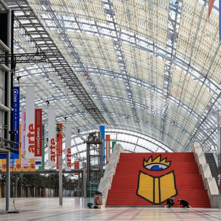 Eine Werbetechnikerin und ein Werbetechniker kleben das Logo der Leipziger Buchmesse auf eine Treppe in der Glashalle der Leipziger Messe. 