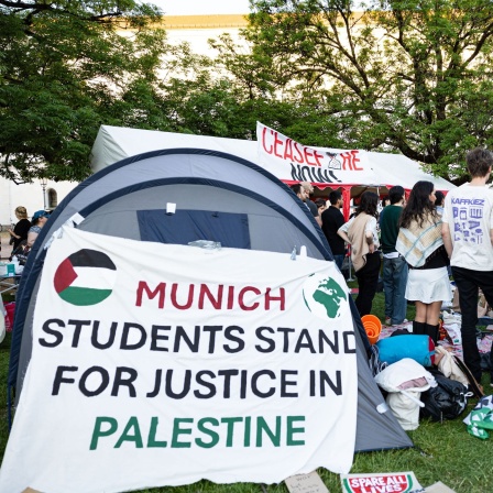 Studierende stehen zwischen Zelten. An einem davon klebt ein Banner mit der Aufschrift "Munich students stand for justice in Palestine"