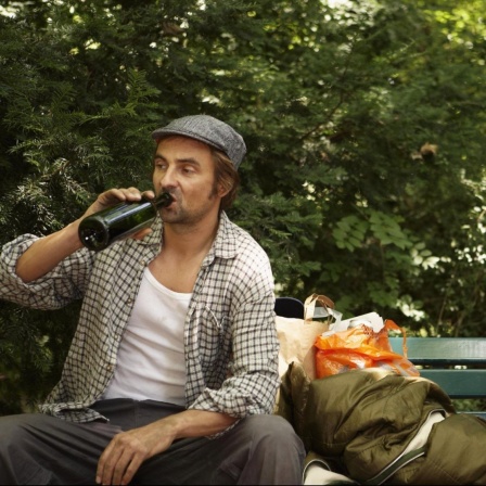 Ein Mann sitzt mit einem Bier und großer Tasche auf der Bank, ein anderer im Anzug schaut ihn spektisch an.