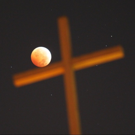 Ein religiöses Kreuz vor dem Nachhimmel, während einer totalen Mondfinsternis. Der rot erleuchtete Mond scheint das Kreuz zu berühren.