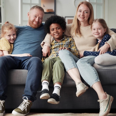 Eine Familie sitzt gutgelaunt zusammen auf dem Sofa, Mutter, Vater und drei Kinder