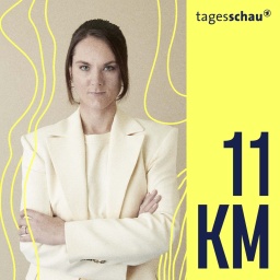 11KM-Host Victoria Koopmann steht im weißen Anzug  mit verschränkten Armen vor einer beigen Wand. 