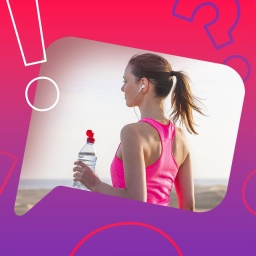 Eine junge blonde Frau in Sportkleidung und mit Kopfhörern steht mit einer Getränkeflasche in der Hand an einem Strand